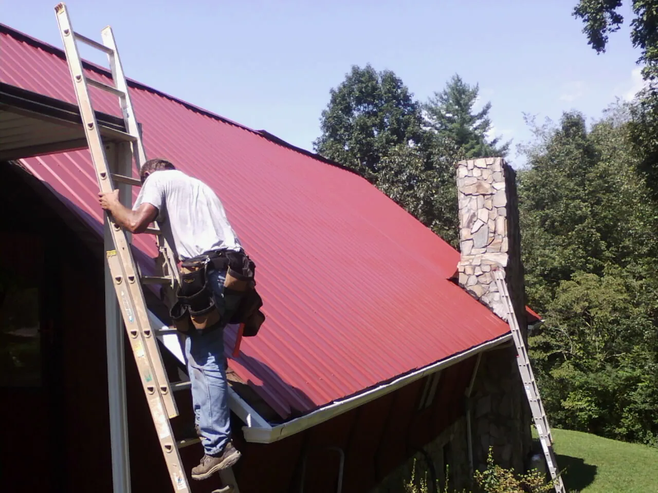 Roofer on a ladder beside a red metal roof and stone chimney — Soffit & Fascia Repair by Roofing Brothers in Woods Cross
